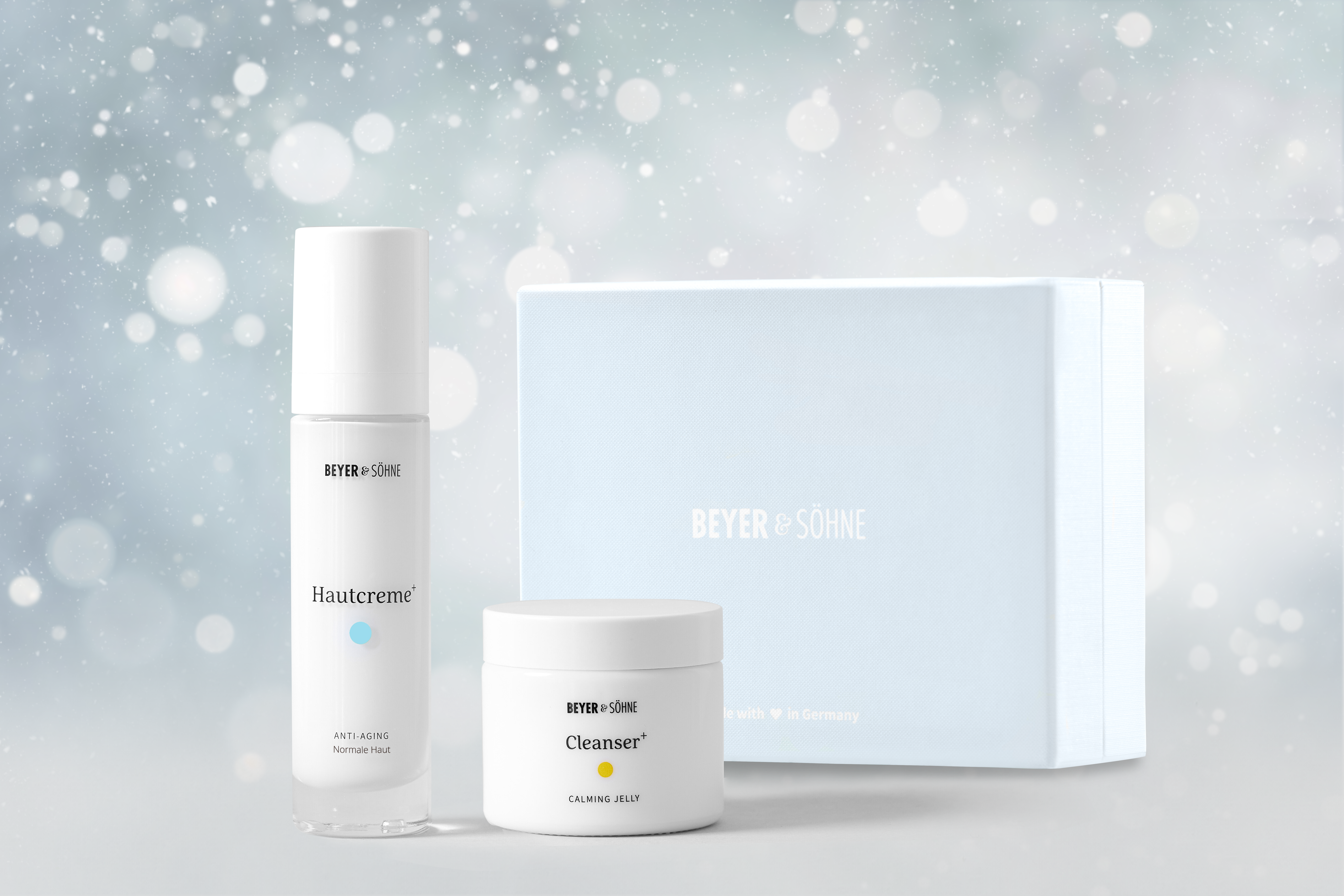 Hautcreme+ für normale Haut und Cleanser+ von Beyer & Söhne in hochwertigem Glasspender und Tiegel vor hellblauer Geschenkbox und weihnachtlich gestimmten Hintergrund mit Schneeflocken.